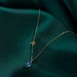 14K gold necklace with bird charm and blue sapphire teardrop pendant on emerald green satin fabric