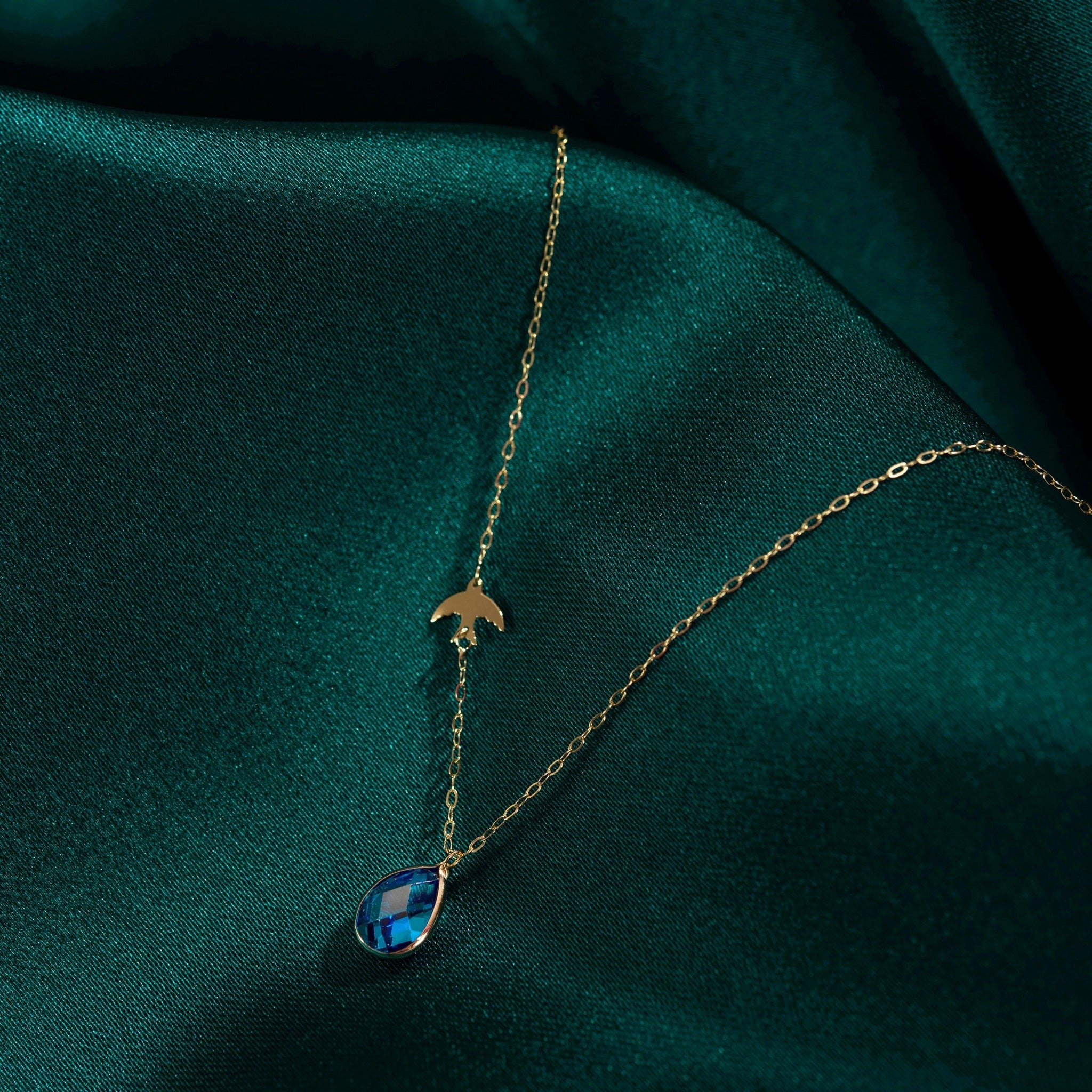 14K gold necklace with bird charm and blue sapphire teardrop pendant on emerald green satin fabric