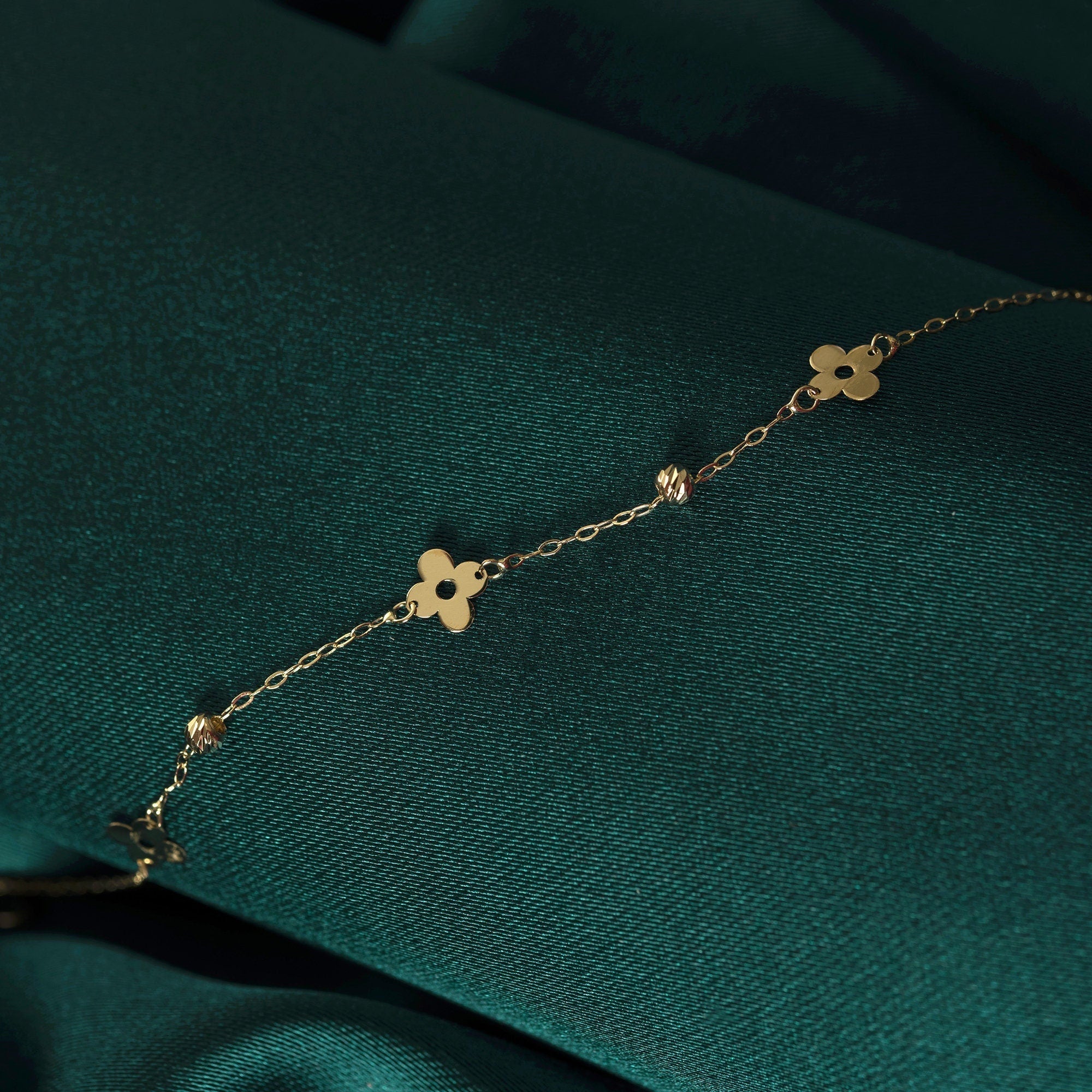 Minimalist 14K gold bracelet with clover-shaped charms and tiny textured accents, styled on emerald green silk fabric