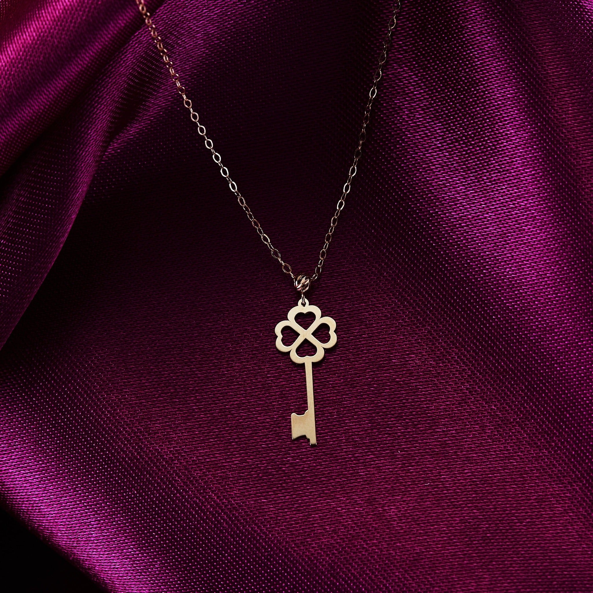 14K gold necklace with a key-shaped pendant featuring a four-leaf clover design, styled on magenta satin fabric.