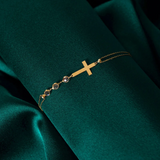 14K gold bracelet with minimalist cross and three small crystal beads, displayed on dark green satin fabric