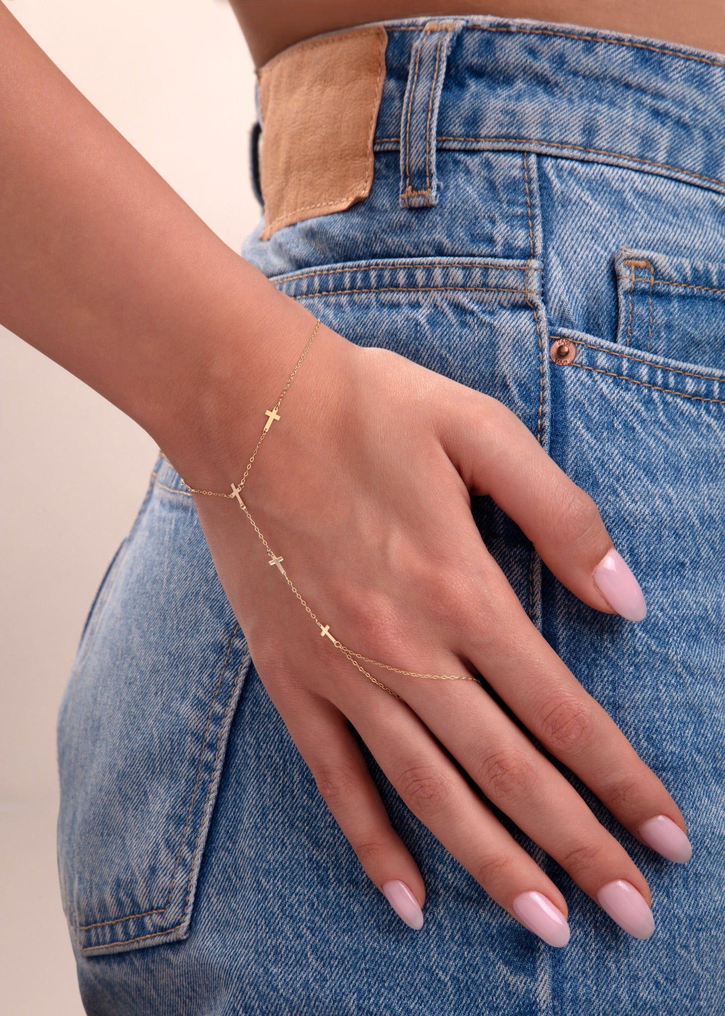 Model wearing a 14k gold hand chain bracelet with multiple cross charms over blue denim pants

