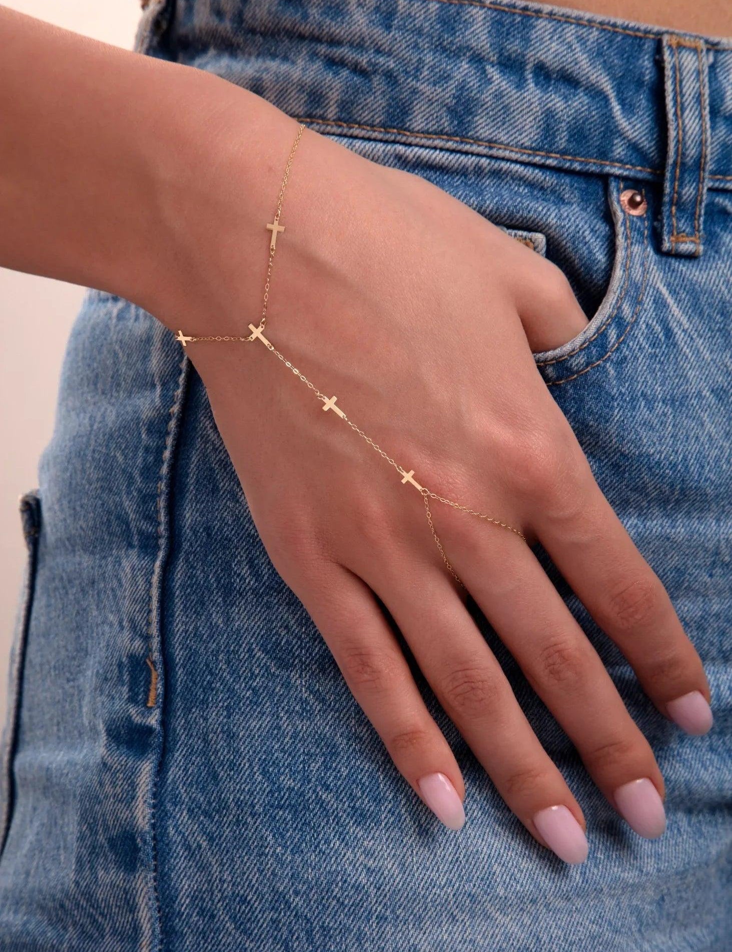Close-up of a woman wearing a 14k gold hand chain bracelet with cross charms, styled with blue jeans

