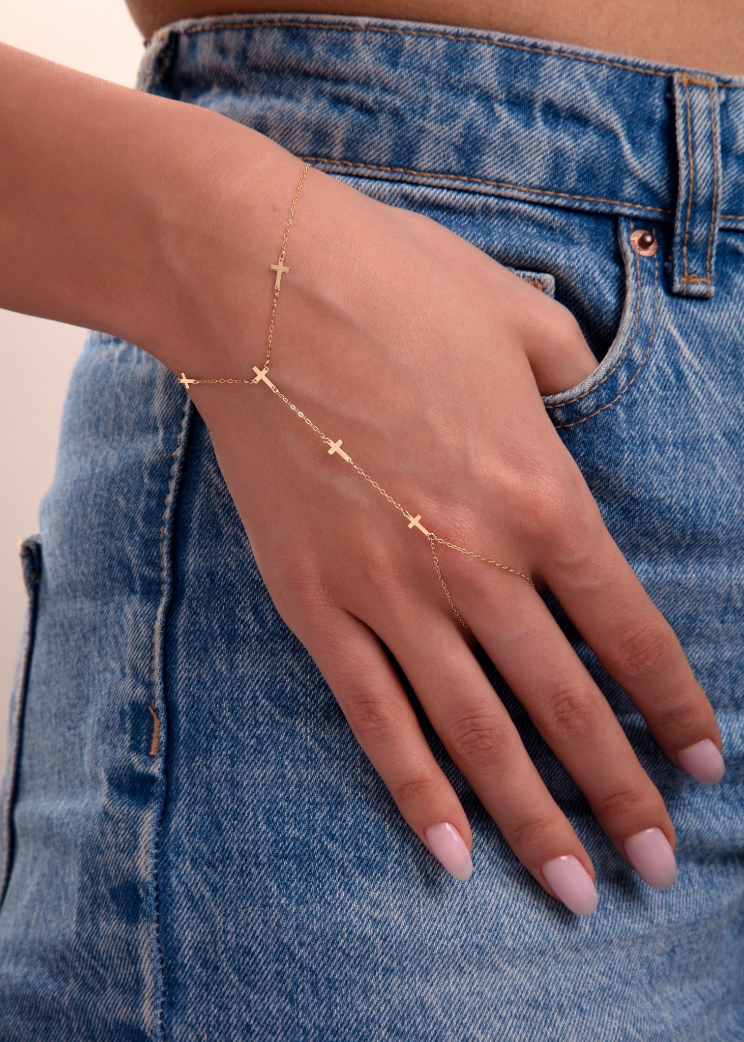 Close-up of a woman wearing a 14k gold hand chain bracelet with cross charms, styled with blue jeans

