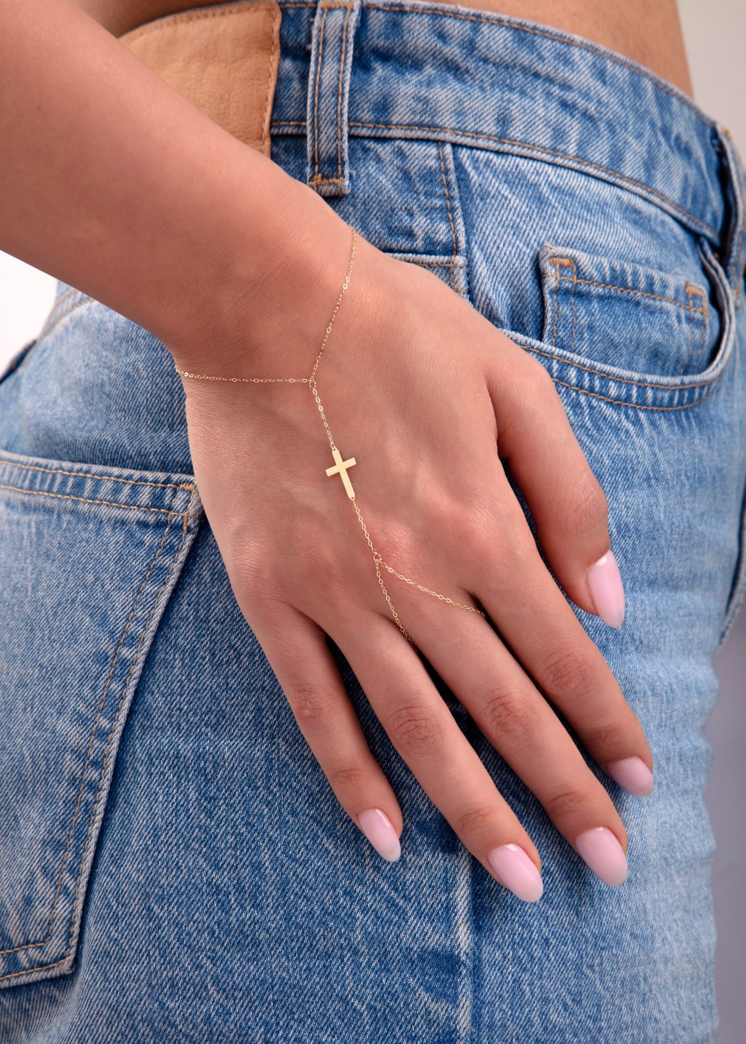 Close-up of a 14k gold hand chain bracelet with a single cross charm on a woman's hand in jeans

