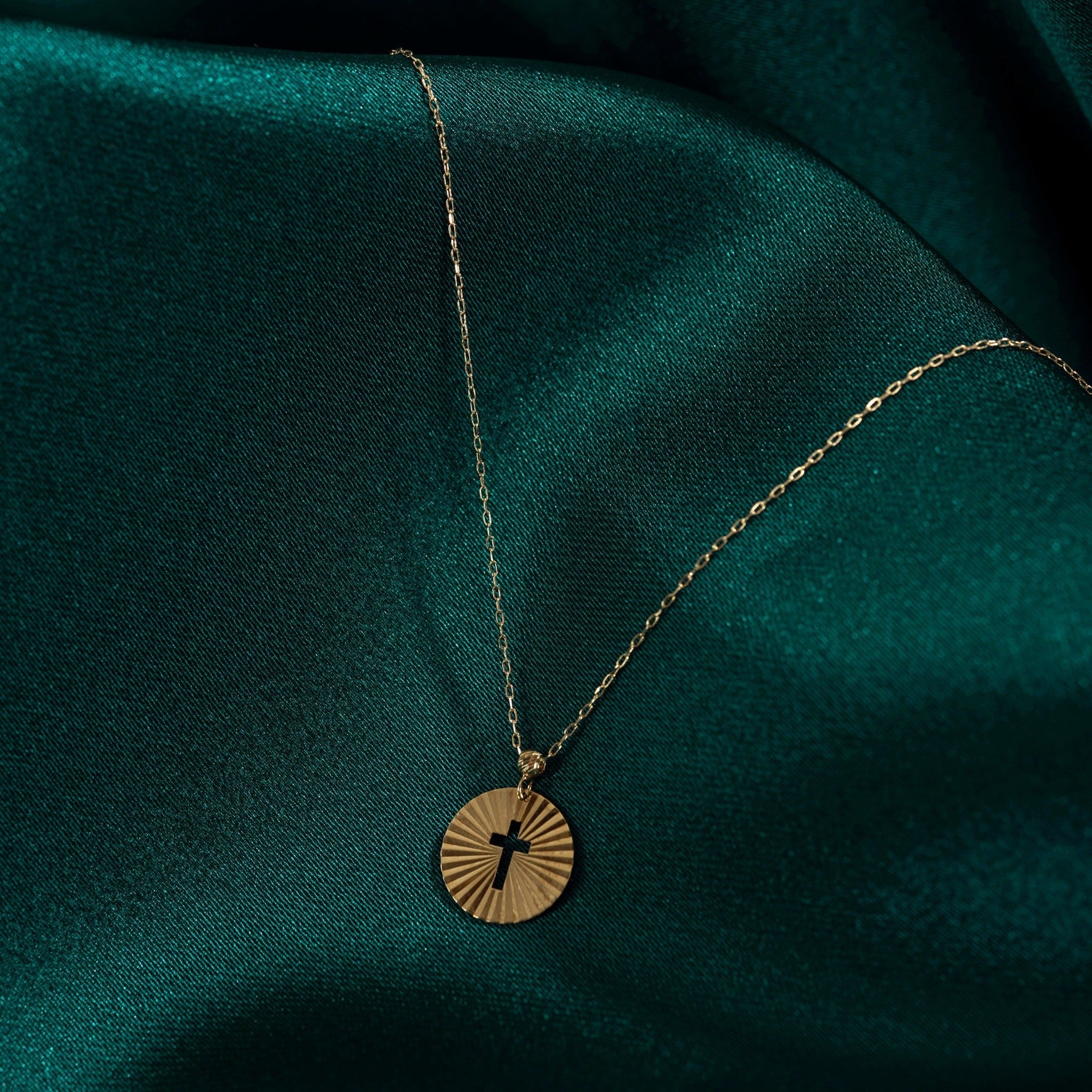 Minimalist 14K gold medallion necklace with a central cutout cross and radiant sunbeam engravings, styled on emerald green satin.