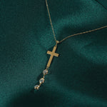 Minimal 14K gold cross necklace with crystal drop accents photographed on emerald green silk background
