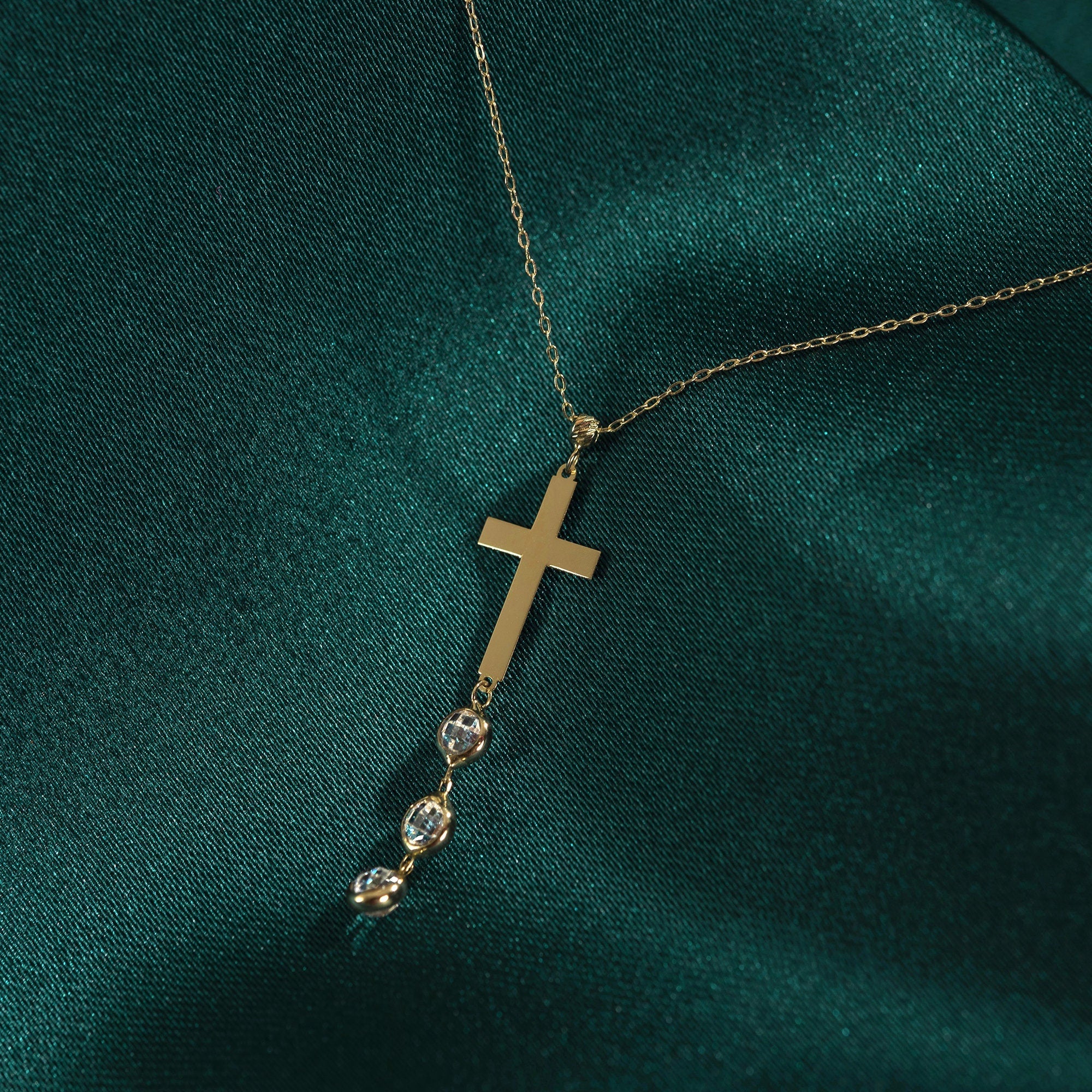 Minimal 14K gold cross necklace with crystal drop accents photographed on emerald green silk background