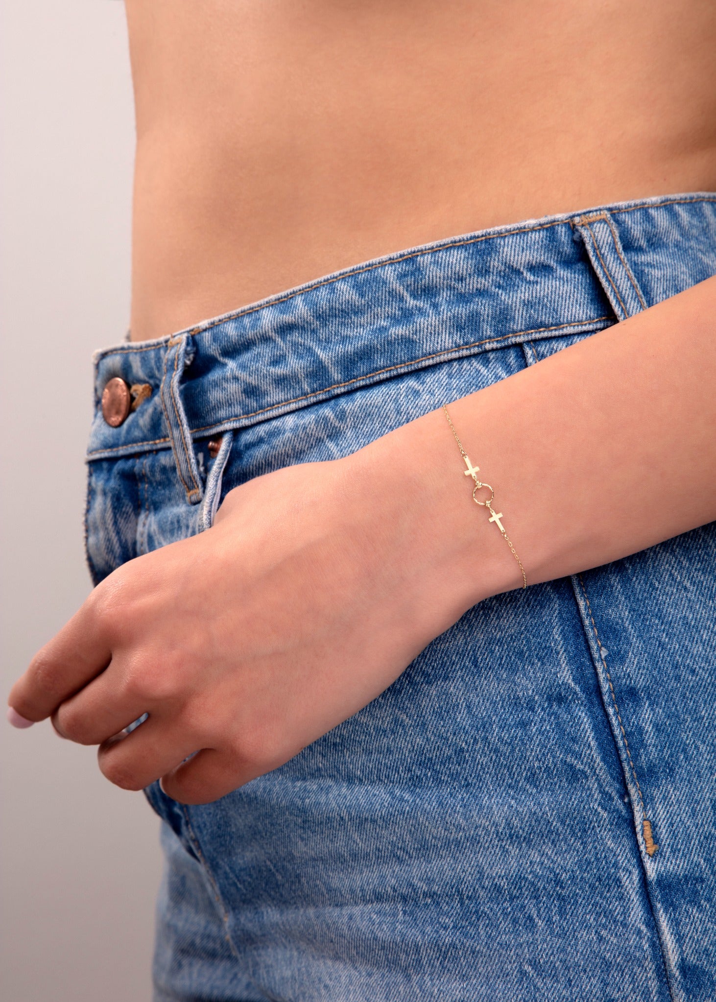Elegant 14k gold bracelet with cross and circle charms on a model’s wrist wearing blue jeans

