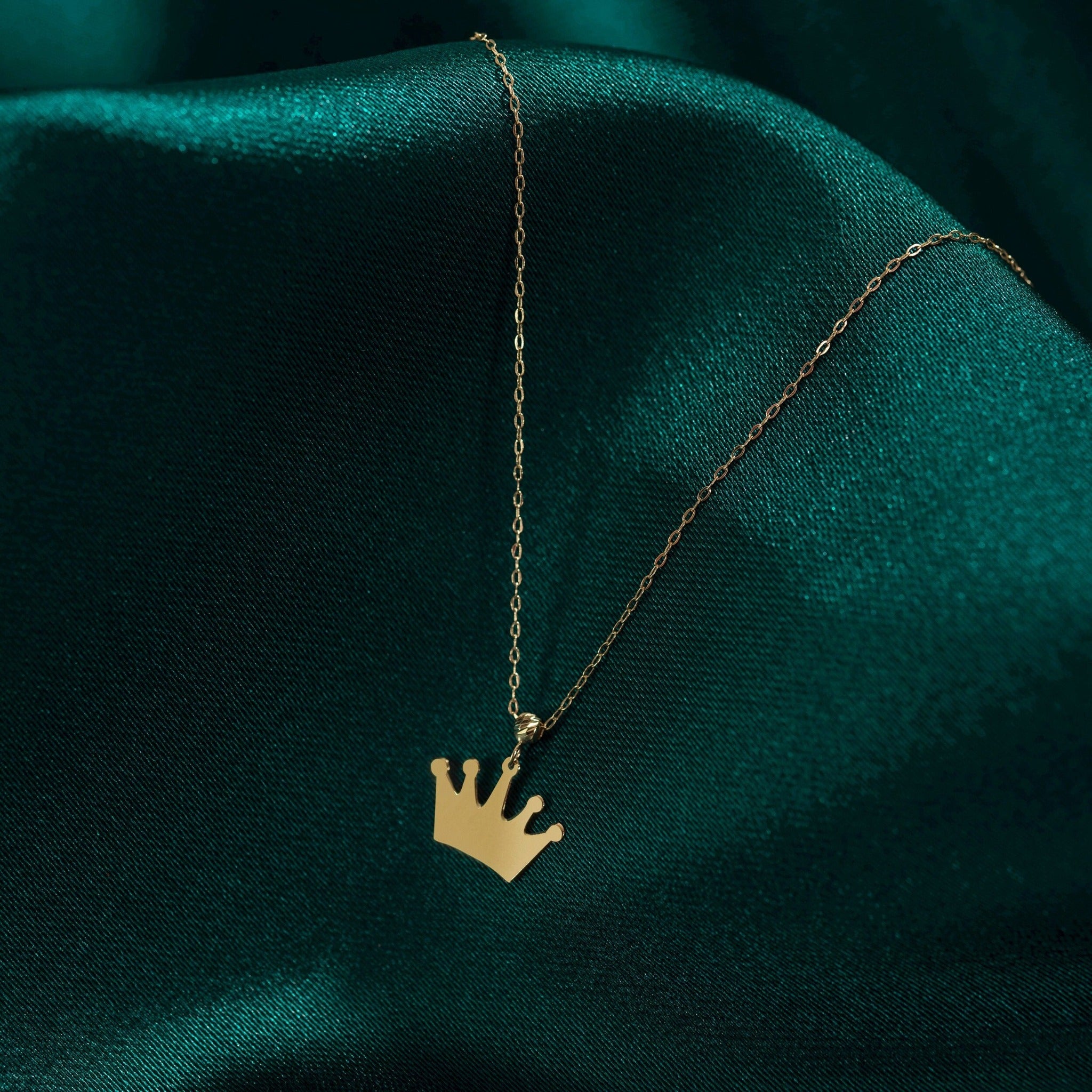14K gold necklace with a polished crown-shaped pendant, resting on a deep green fabric background – simple and elegant royal jewelry.