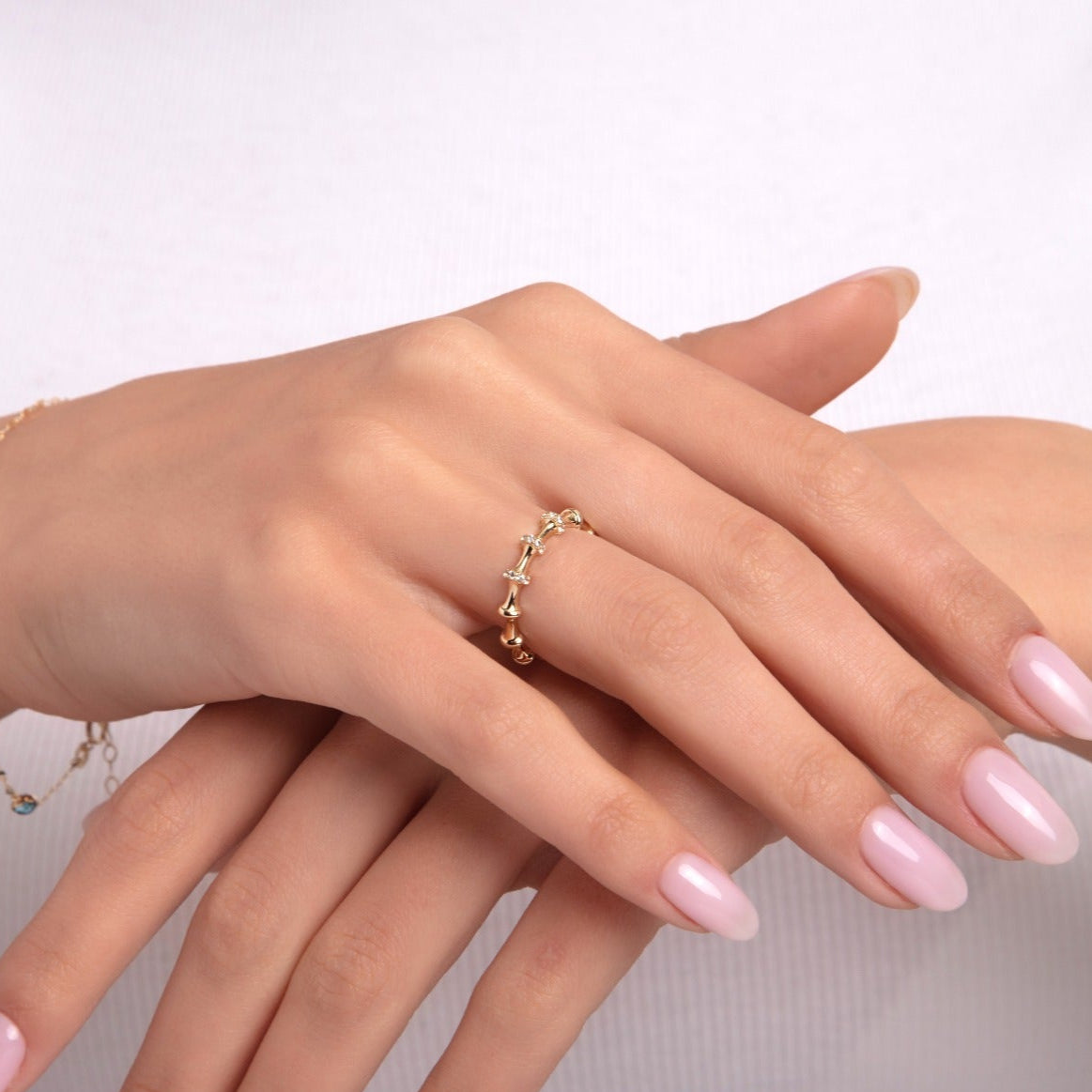 Delicate 14K gold ring with tiny bell-like charm drops, shown on a woman's hand with light pink nails and a white long sleeve top, styled with a minimal beaded bracelet.