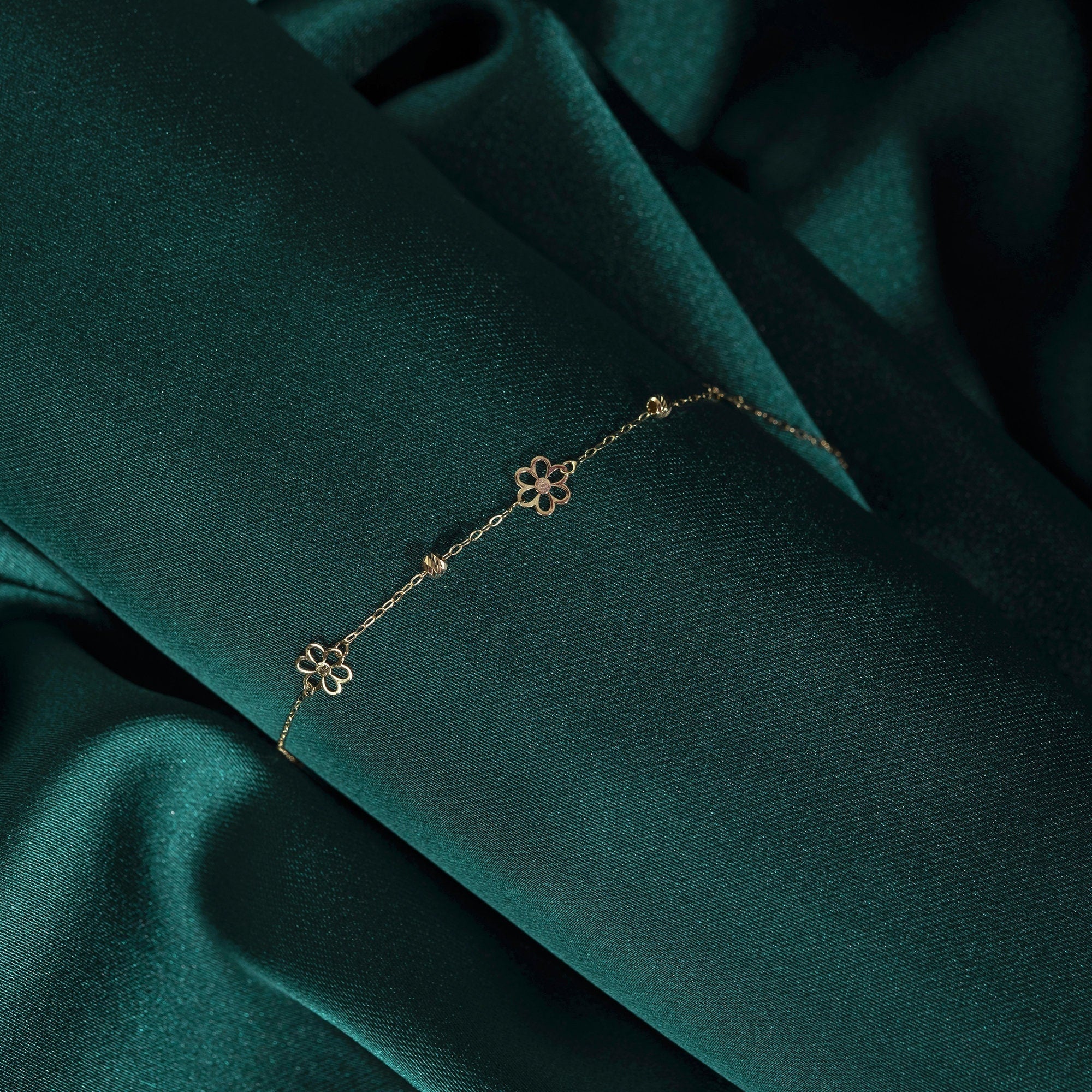 14K gold bracelet featuring three dainty flower charms and bead accents, styled on emerald satin fabric