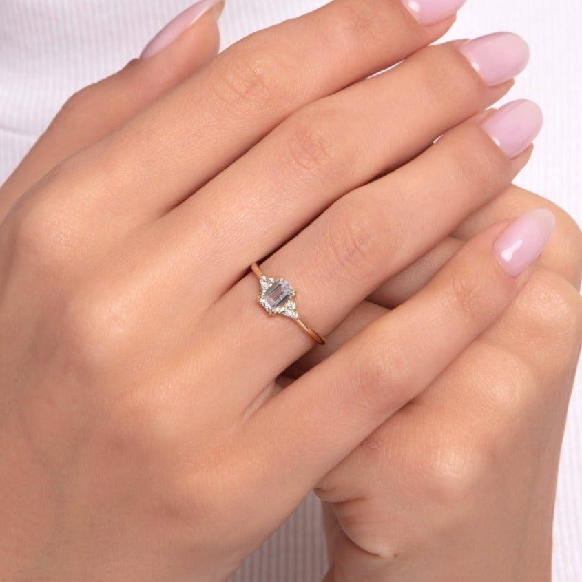 Elegant emerald-cut halo engagement ring with baguette side stones, shown on a woman's hand with soft pink nails and a white turtleneck top.

