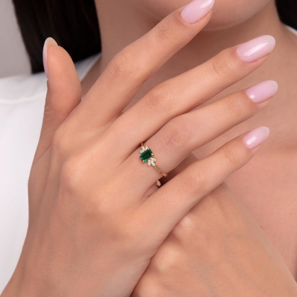 Woman wearing a 14K gold ring with a vivid emerald-cut green gemstone and diamond clusters.
