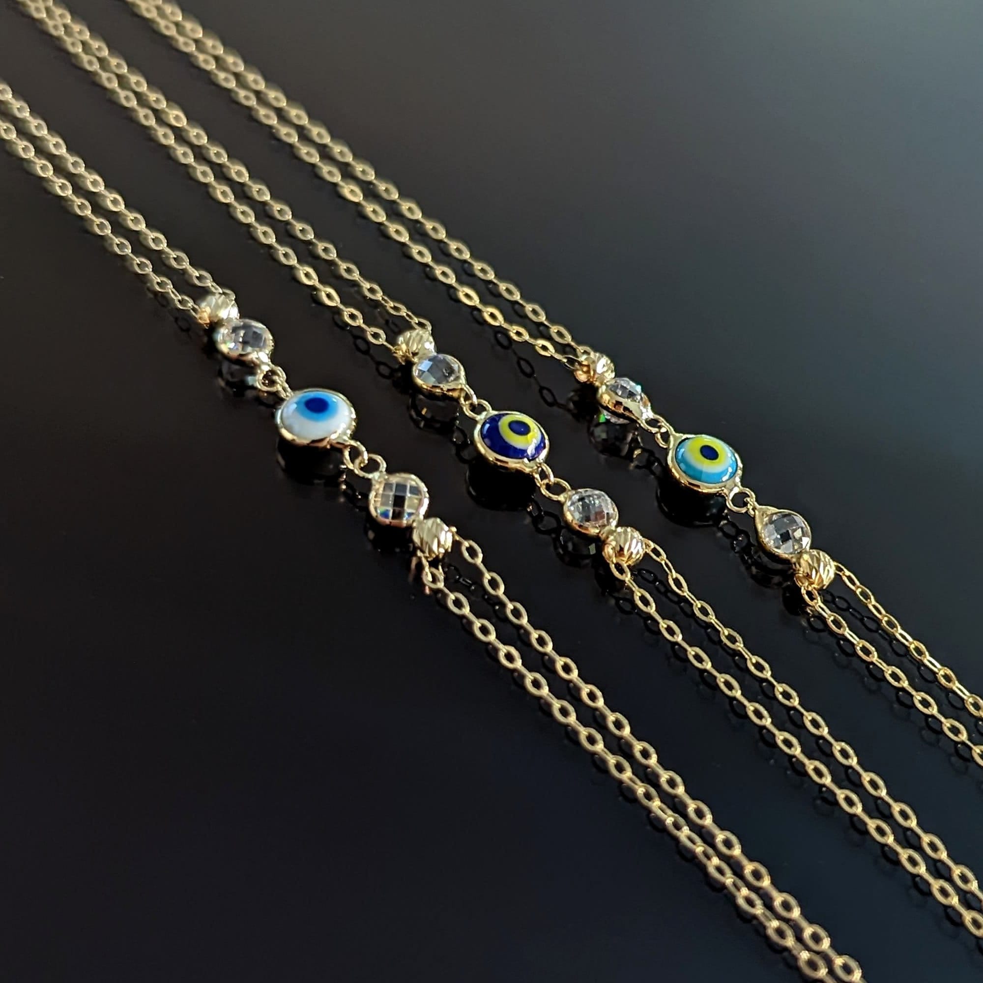 Three 14K gold evil eye bracelets with colorful charms and gemstone accents on black background