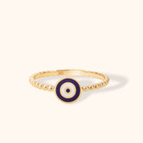 14K yellow gold ring with blue and white enamel evil eye motif on beaded band, photographed on cream background.