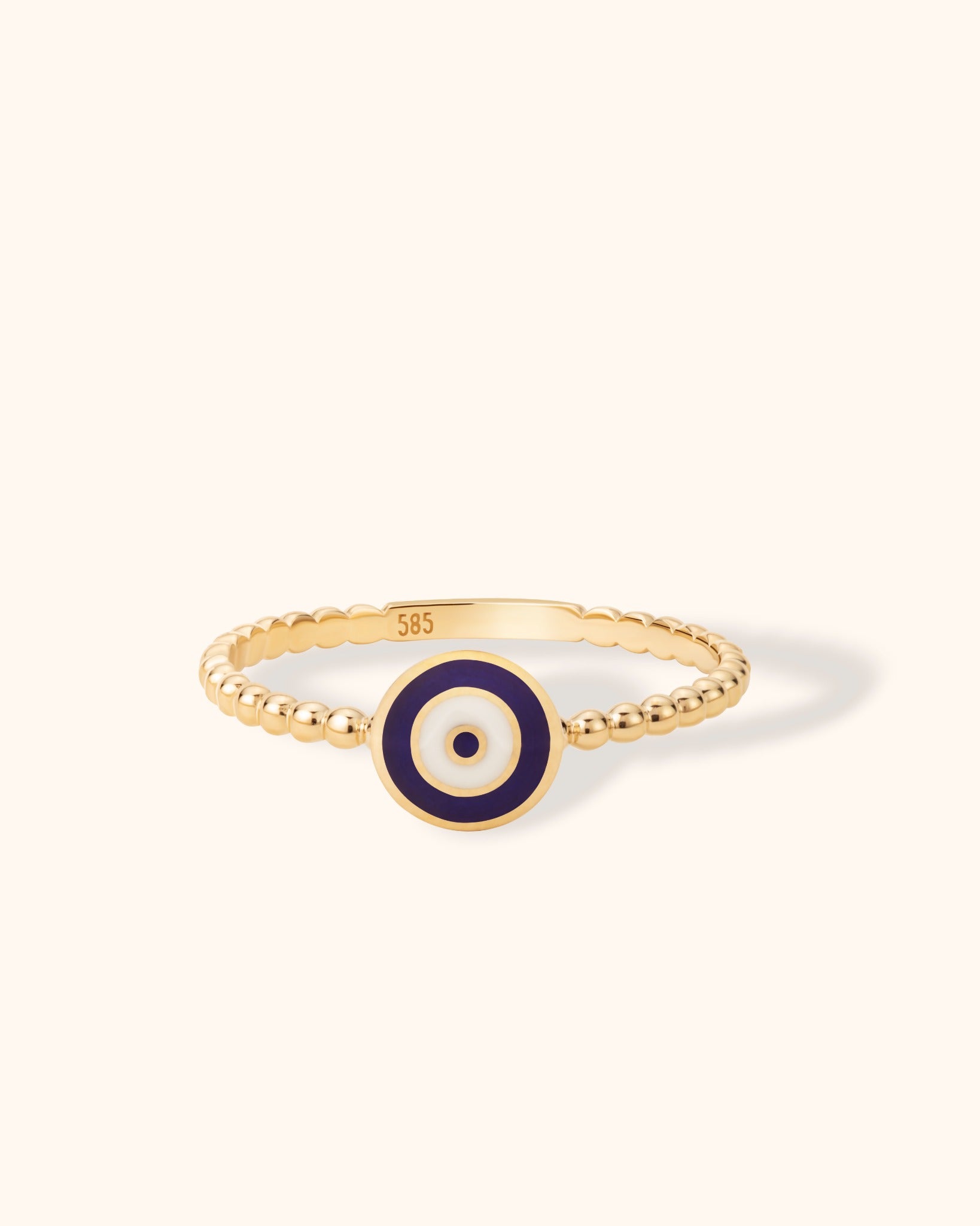 14K yellow gold ring with blue and white enamel evil eye motif on beaded band, photographed on cream background.