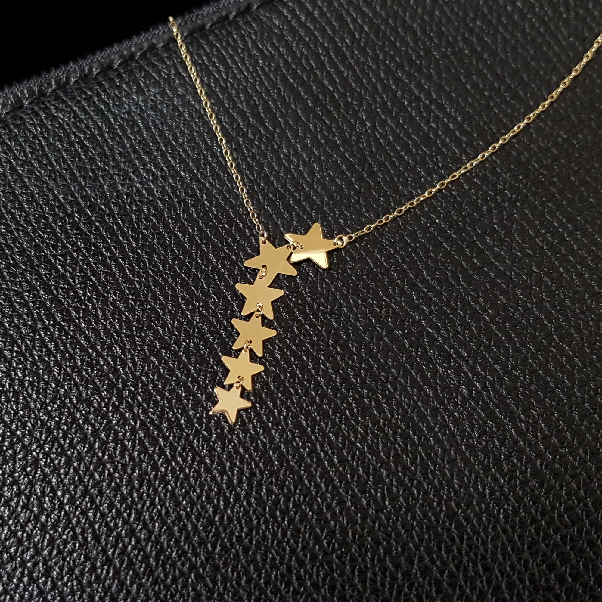 Seven-star cascade necklace in 14K gold displayed on black textured leather background, featuring minimalist celestial design.