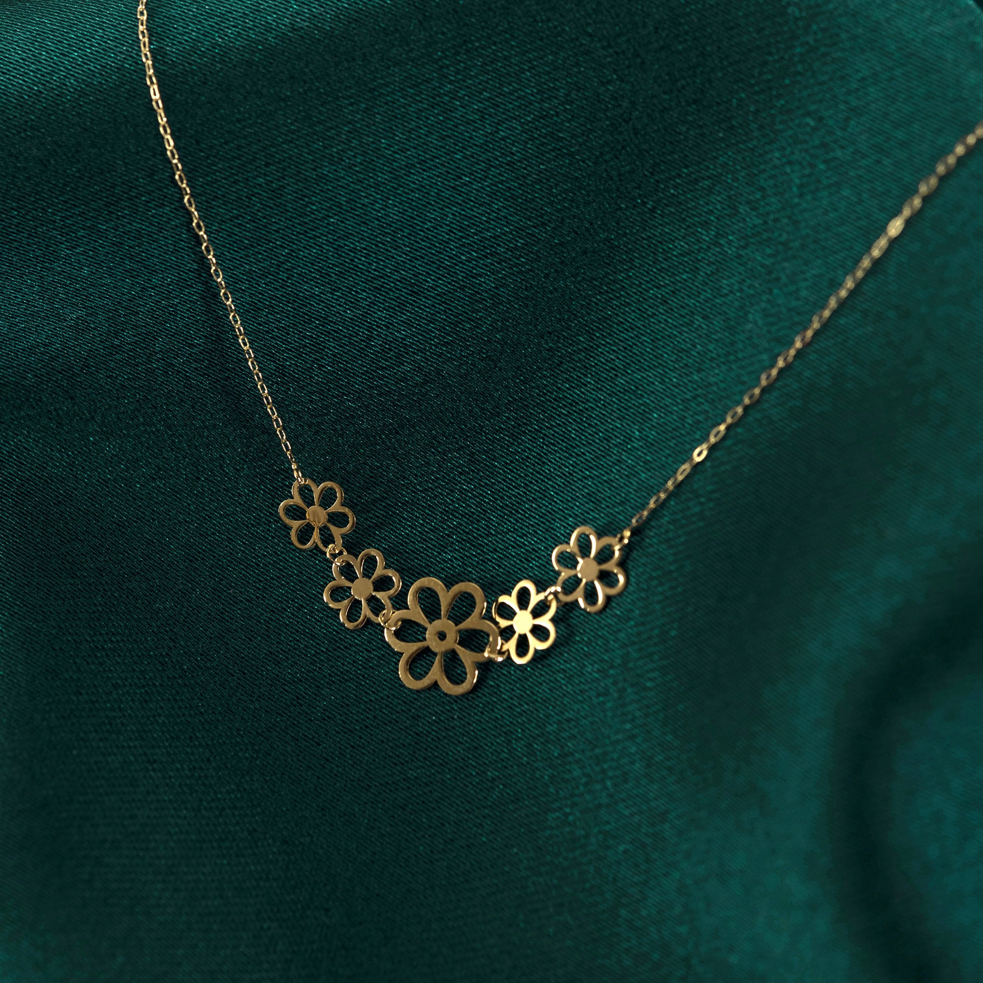 14K gold necklace with five small open flower charms displayed on emerald green satin fabric – elegant and minimalist.