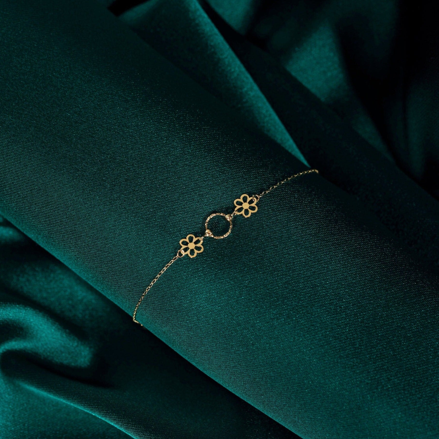 Delicate 14K gold bracelet with two floral charms and a textured gold circle centerpiece, displayed on emerald green fabric