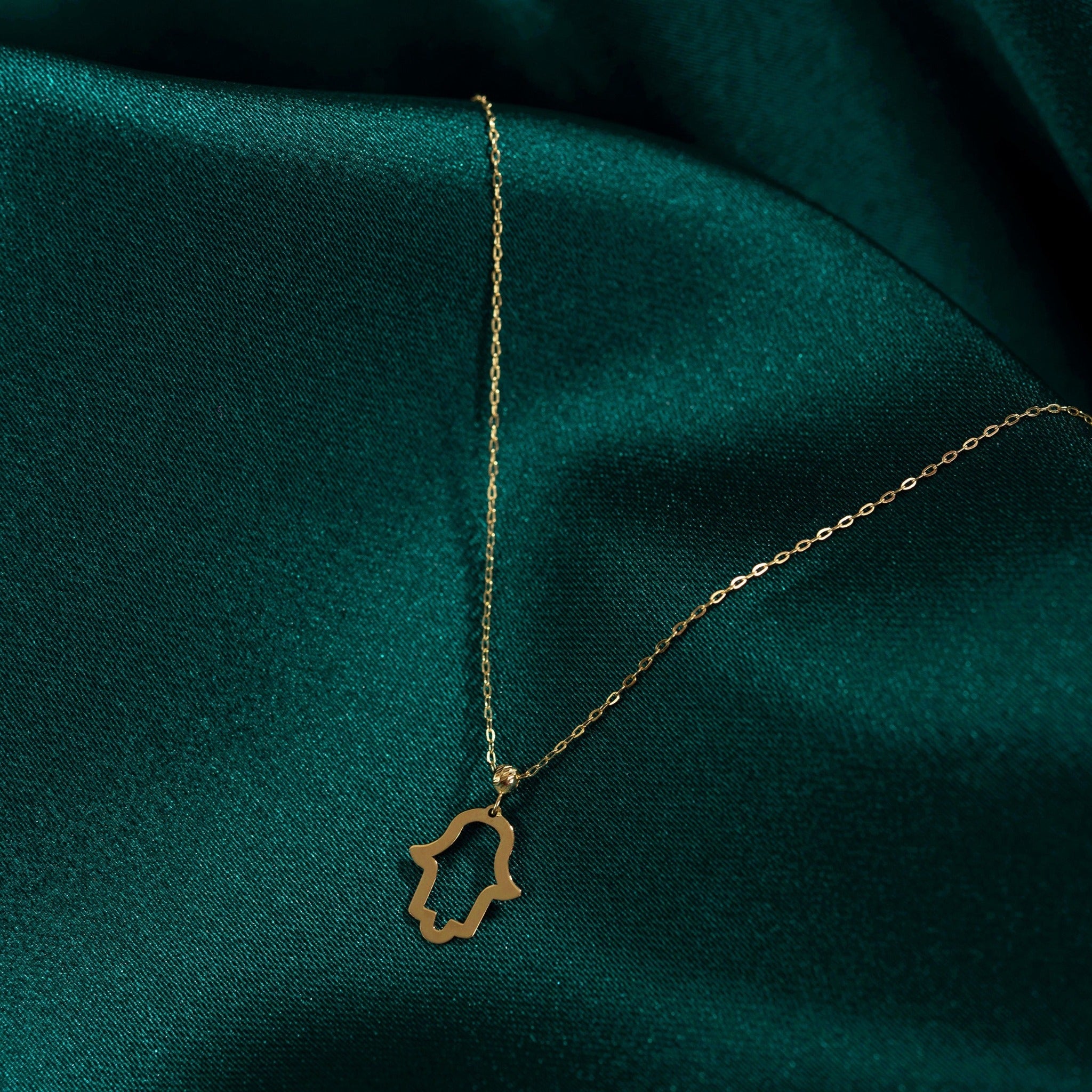14K gold Hamsa hand necklace on emerald green fabric, symbol of protection and spiritual energy