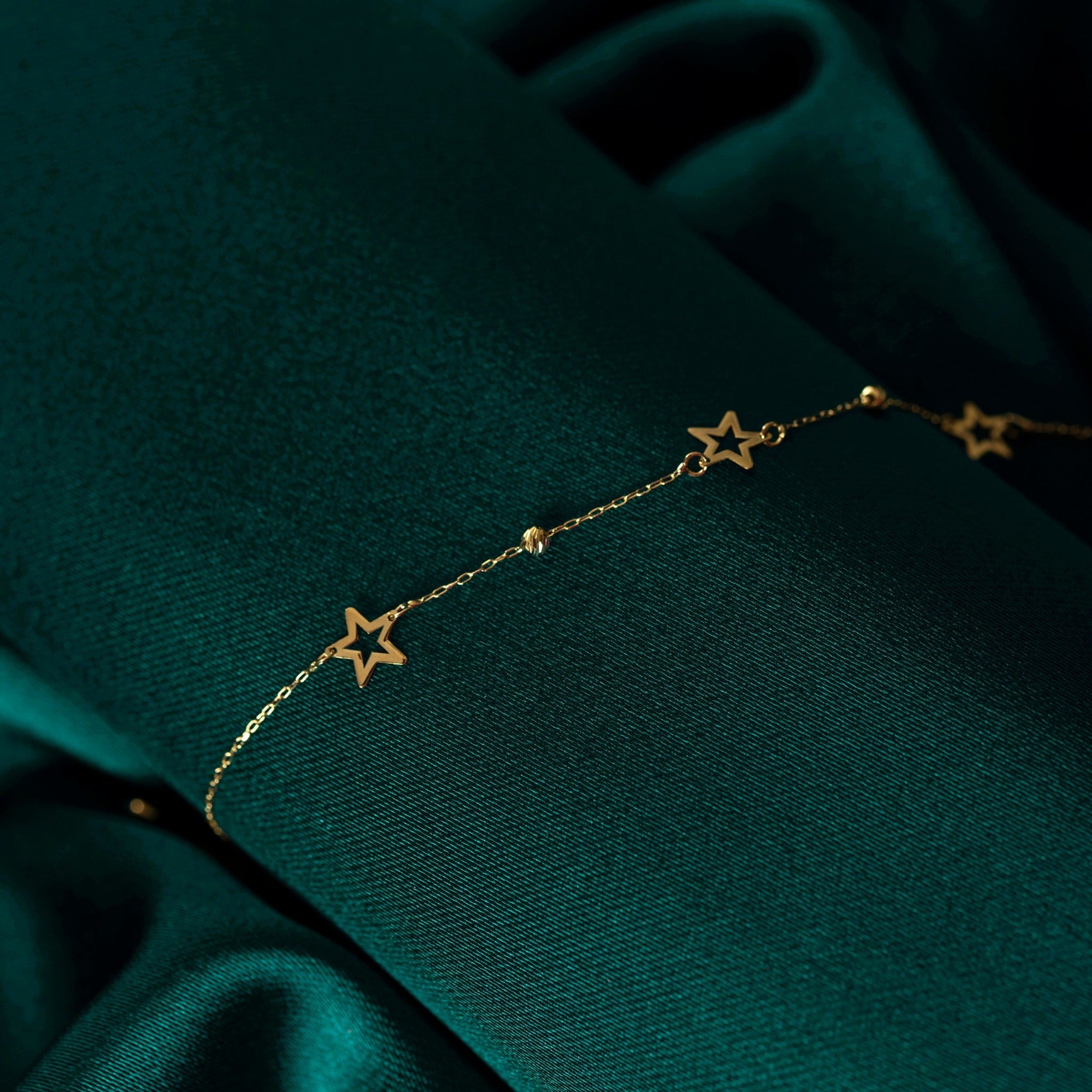 Close-up of a 14K gold bracelet with hollow star charms and gold beads, displayed on dark green satin fabric.