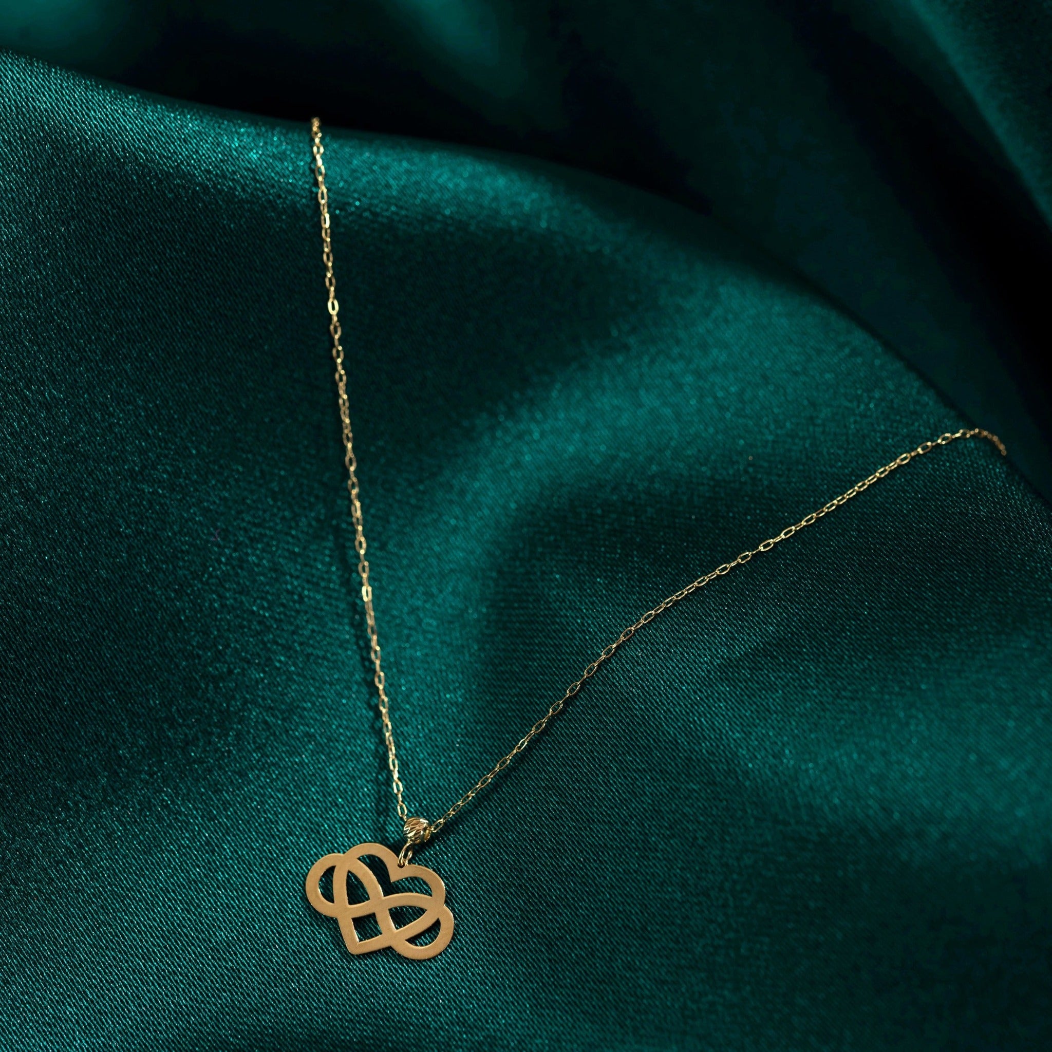 14K gold infinity heart necklace resting on emerald green fabric, symbolizing eternal love and connection.