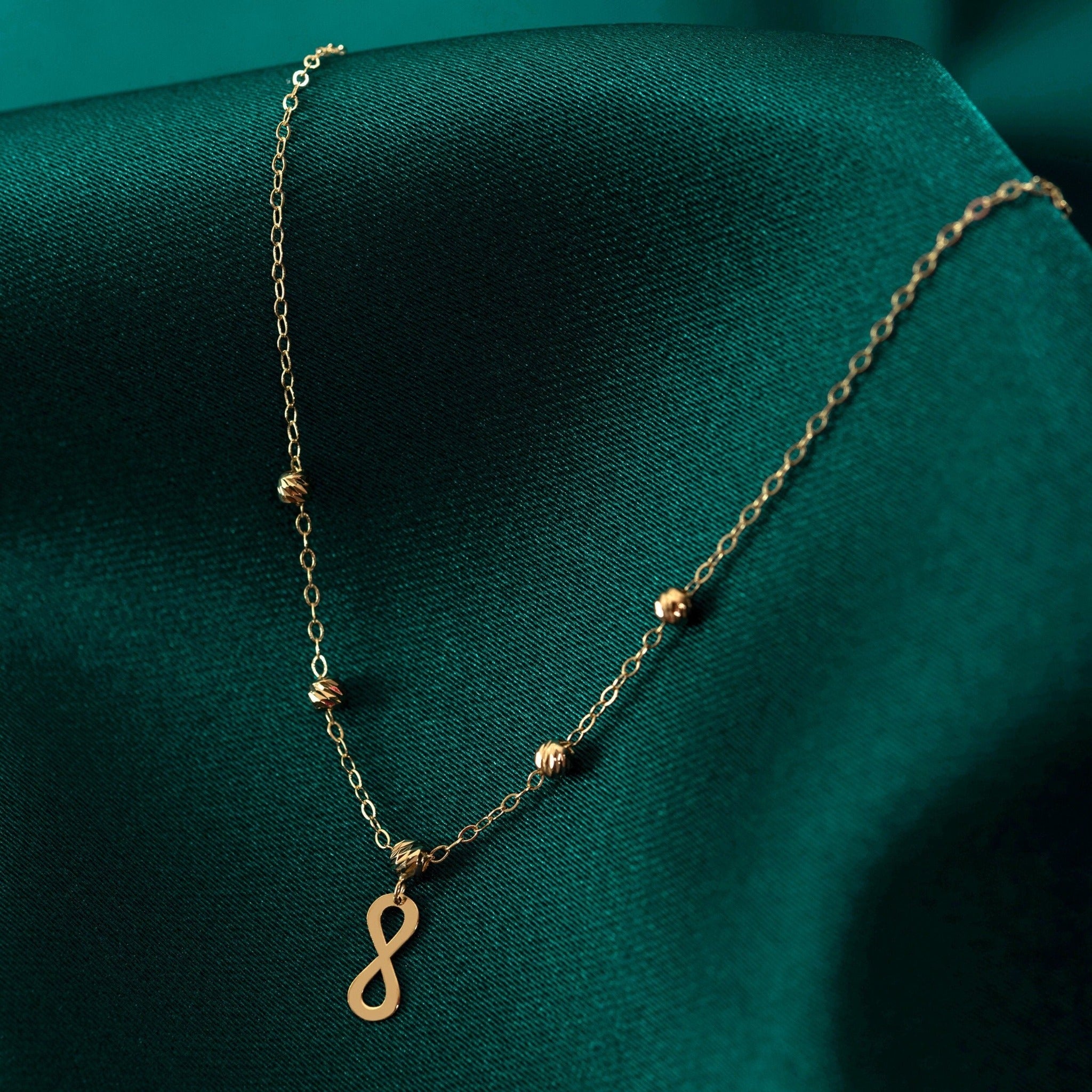Minimalist 14K gold necklace with an infinity pendant and textured beaded chain, displayed on emerald green fabric.