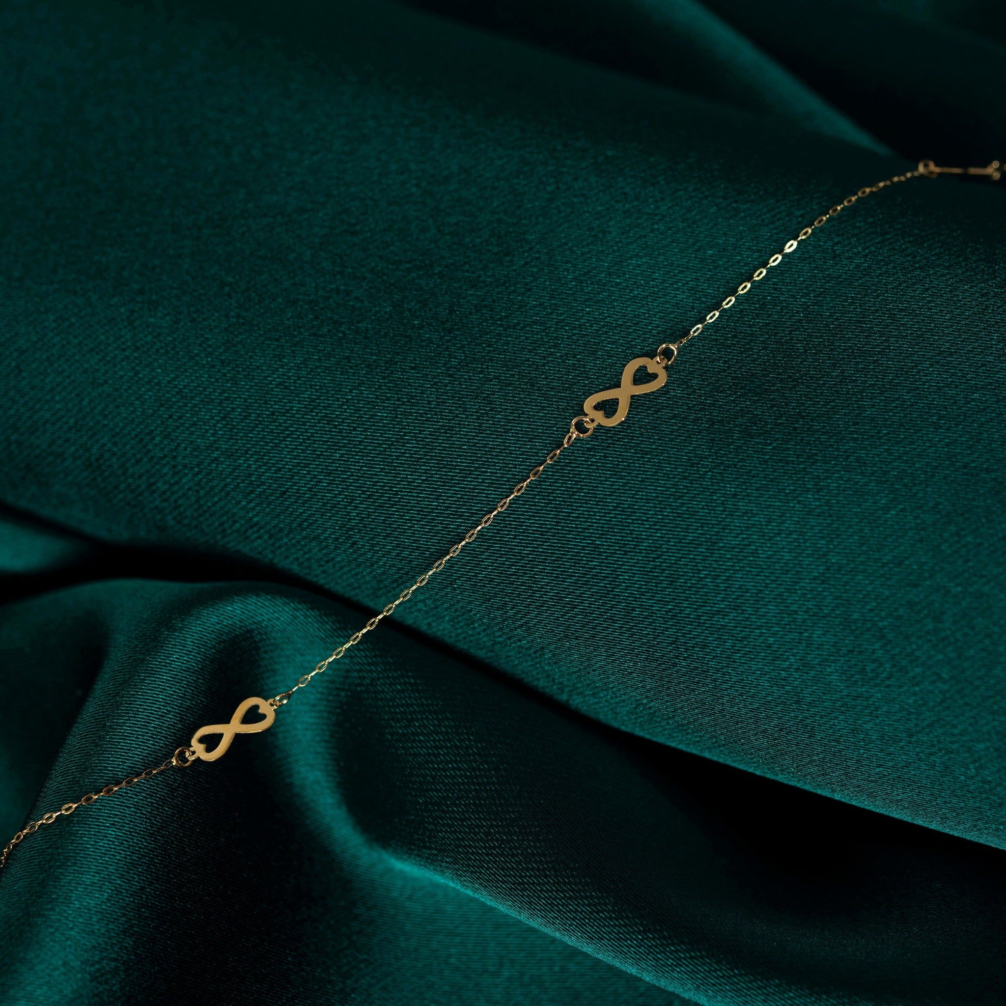 Minimalist 14K gold bracelet with repeating infinity symbols, displayed on emerald green satin