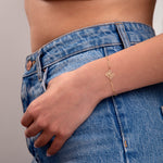 Gold initial bracelet with letter M charm worn on wrist, styled with denim jeans and white knit crop top

