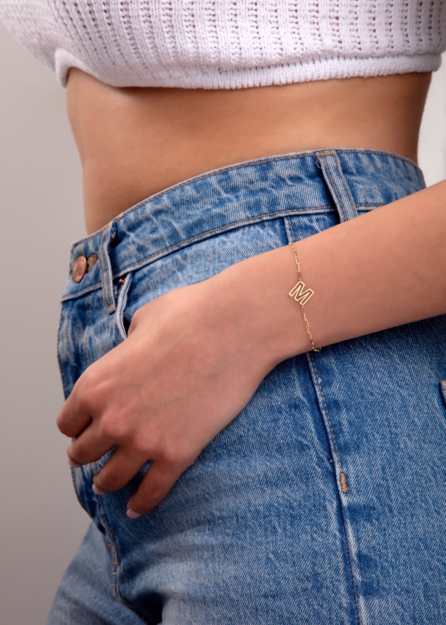 Gold initial bracelet with letter M charm worn on wrist, styled with denim jeans and white knit crop top

