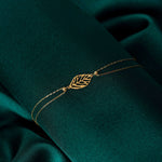 14K gold bracelet with central leaf charm, styled on emerald green satin fabric