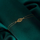 14K gold bracelet with central leaf charm, styled on emerald green satin fabric