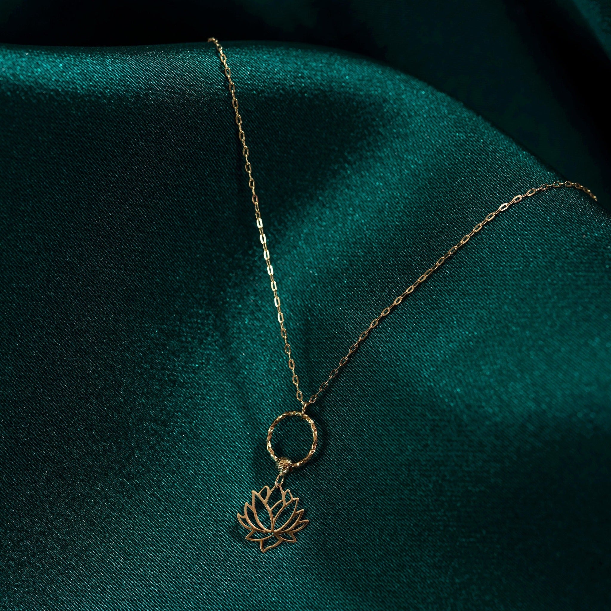 Elegant 14K gold necklace featuring a textured circle and a lotus flower pendant, styled on dark emerald fabric.