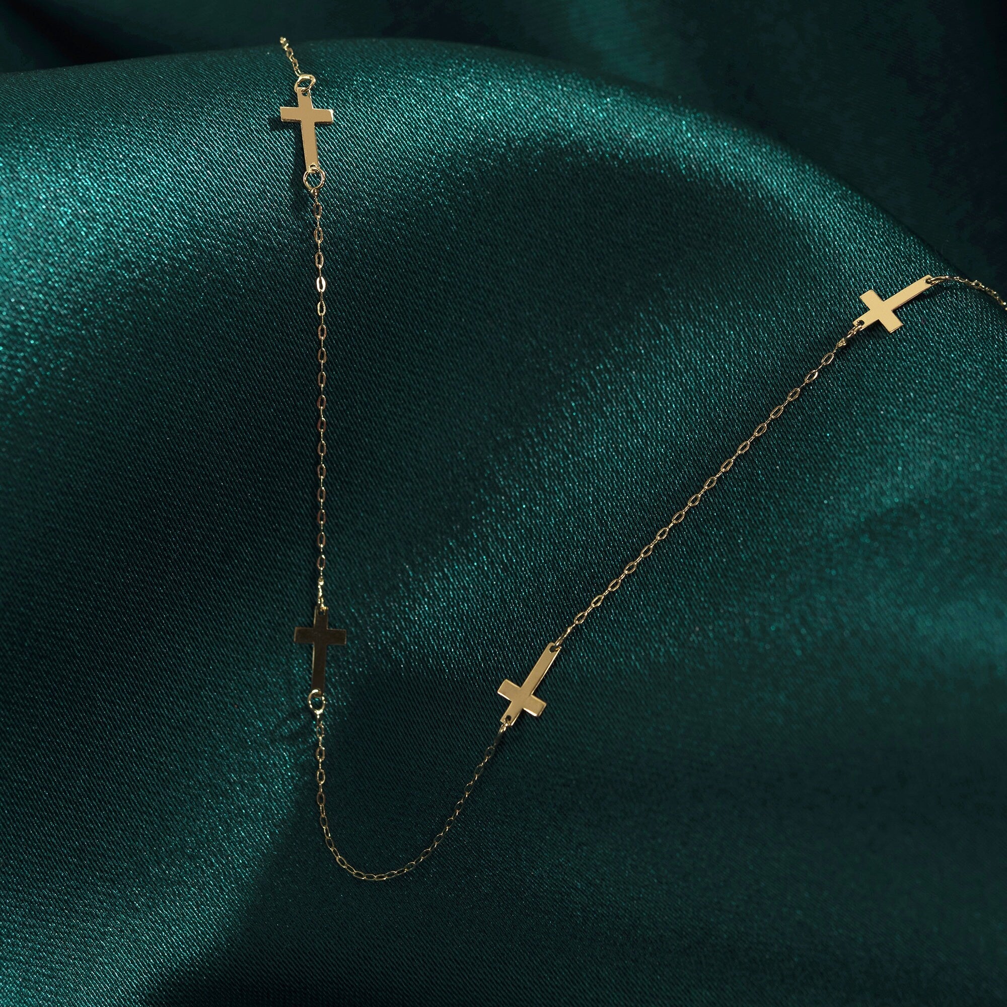 14K gold necklace with multiple small cross charms displayed on dark green satin fabric, elegant and minimalistic.