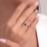 Model's hand showcasing a 14K gold ring with a deep blue sapphire surrounded by halo-set white stones, styled with soft pink nails.