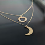 Layered 14K gold necklace with a polished crescent moon and cut-out sun pendant displayed on a dark reflective background.