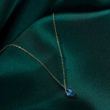 14K gold necklace with a faceted blue teardrop gemstone pendant resting on dark green silk