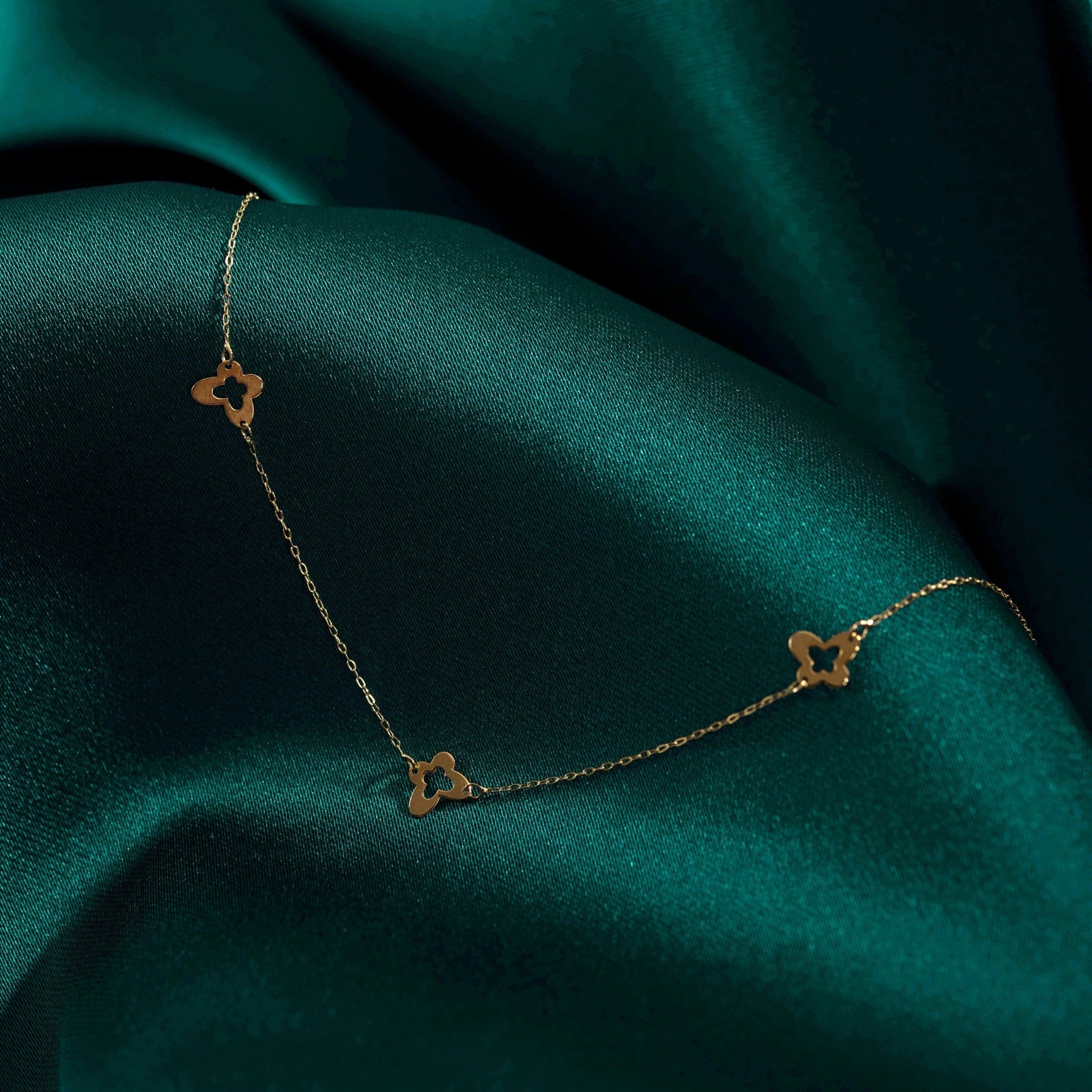 Minimalist 14K gold necklace with three evenly spaced clover charms, set on emerald green satin fabric for an elegant look.