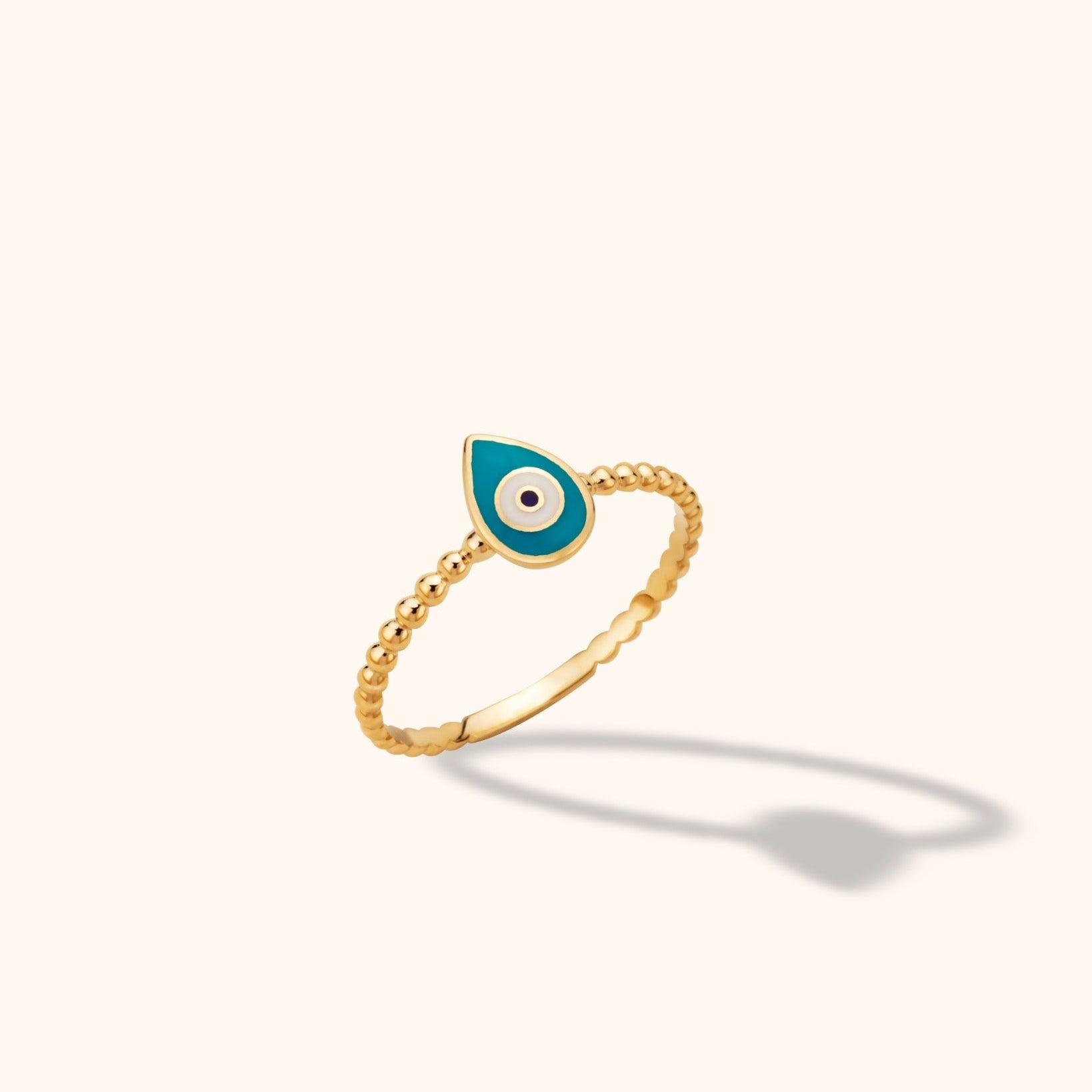 Close-up of a delicate 14K yellow gold beaded band ring featuring a turquoise enamel evil eye motif against a neutral background.