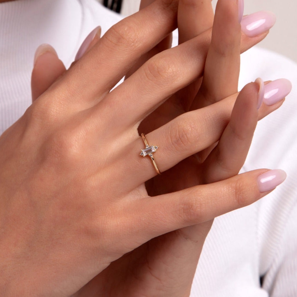 Minimalist 14K gold ring with two baguette-cut diamonds in an open-style setting, worn on a woman’s hand with light pink manicure and white top.