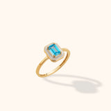 Close-up of a 14K yellow gold ring featuring an emerald-cut blue topaz set within a white enamel octagon, photographed against a neutral background.