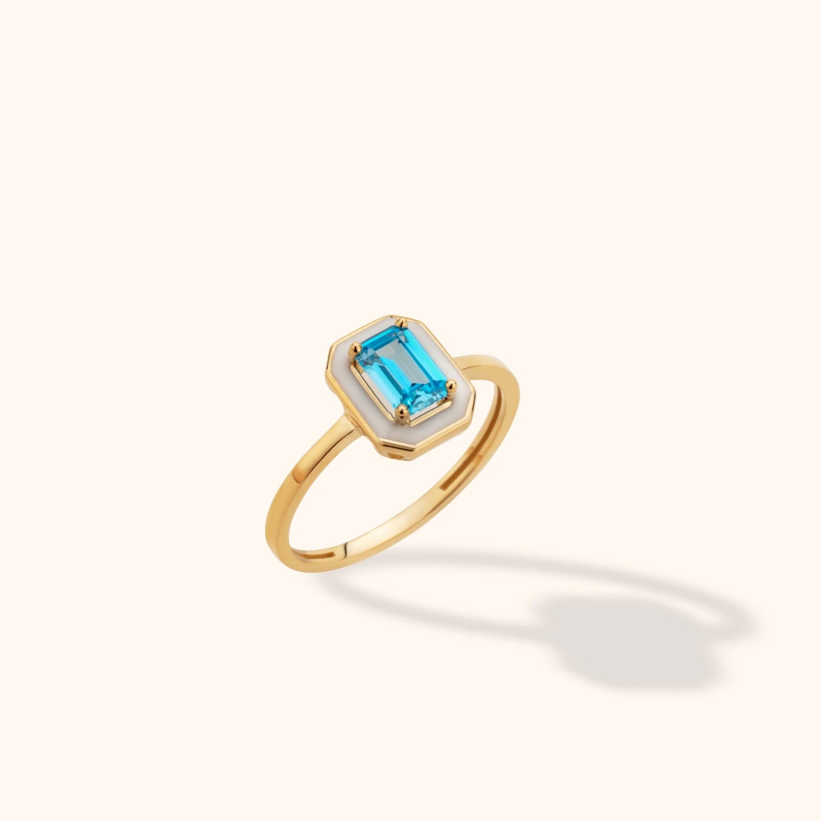 Close-up of a 14K yellow gold ring featuring an emerald-cut blue topaz set within a white enamel octagon, photographed against a neutral background.
