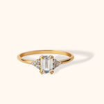 14K yellow gold ring with an emerald-cut center diamond and cluster round diamond accents on each side, shown against a light beige background