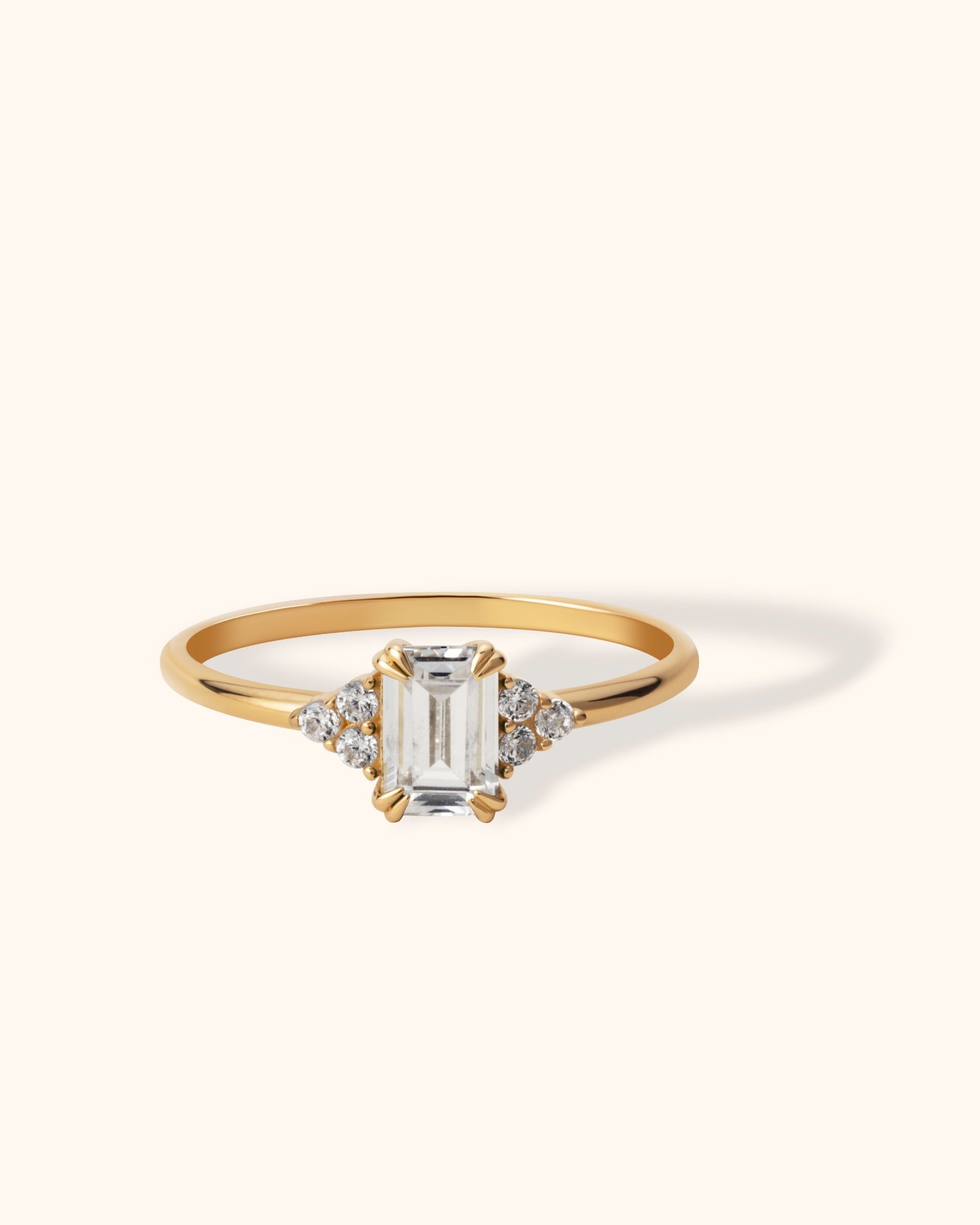 14K yellow gold ring with an emerald-cut center diamond and cluster round diamond accents on each side, shown against a light beige background