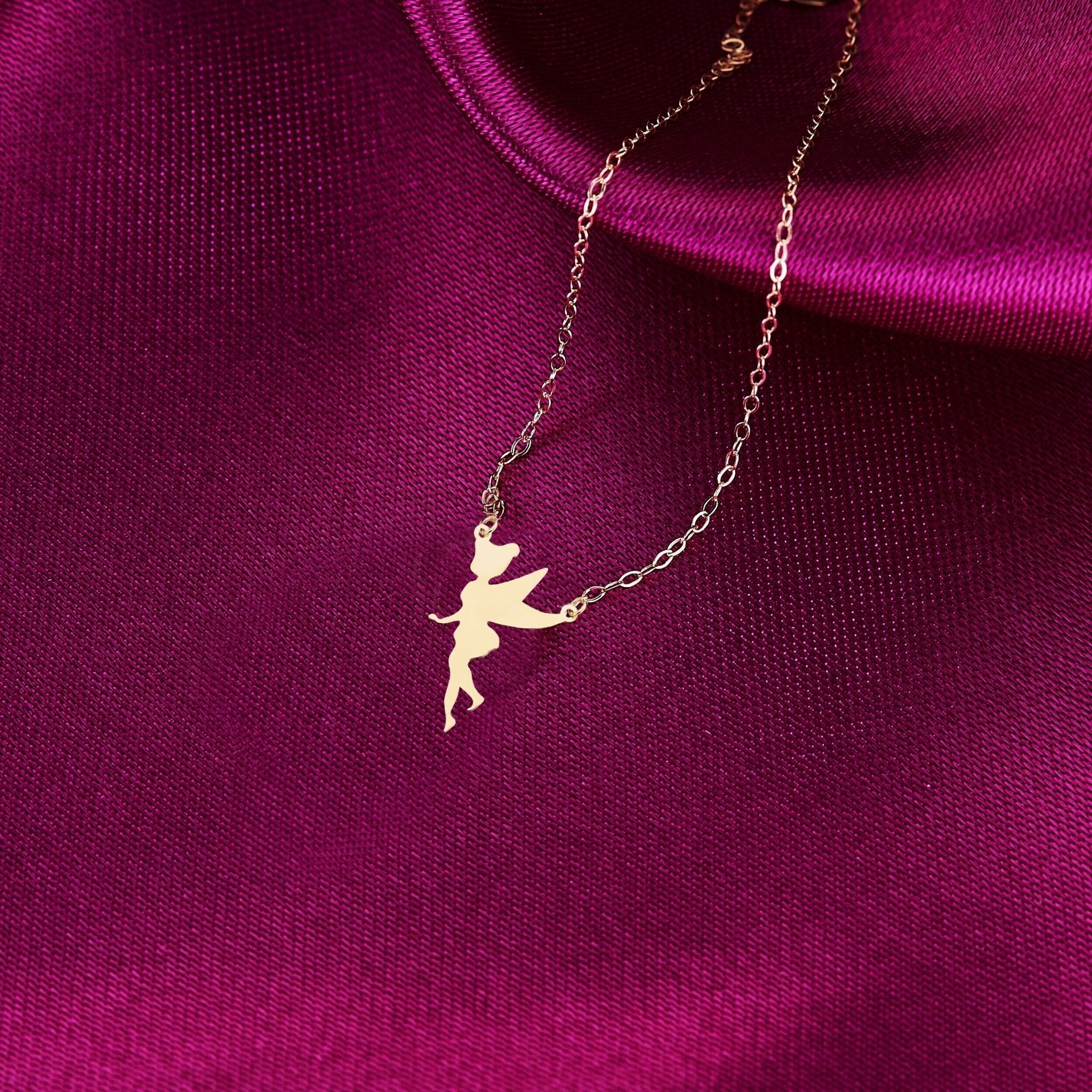 Gold-colored necklace with a flat winged fairy silhouette pendant and fine oval-link chain on deep magenta fabric.