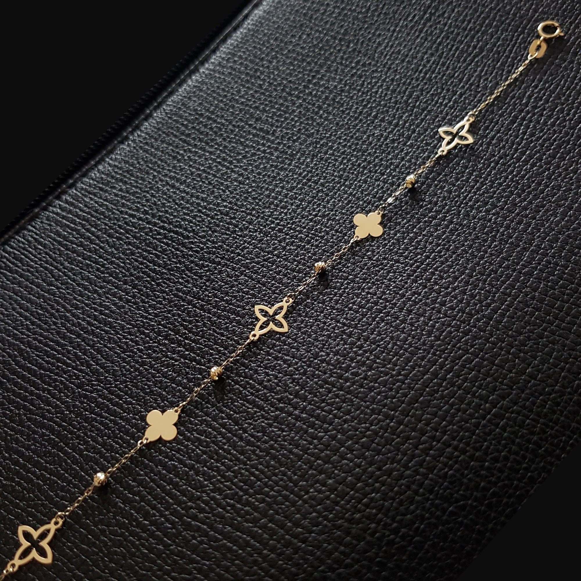 Gold-toned chain bracelet with alternating open and filled clover shapes and small ridged beads on a textured black surface.