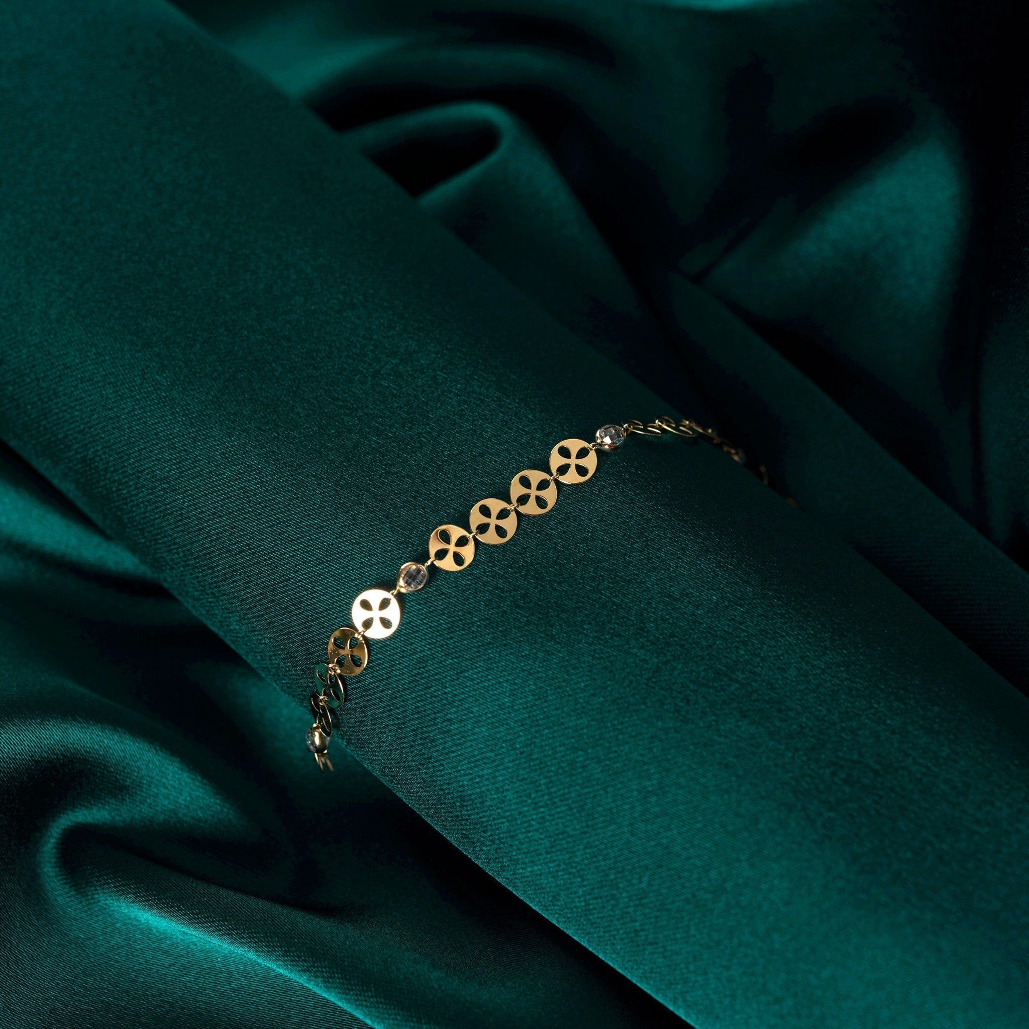 Gold-toned bracelet with round, petal-cutout links and clear faceted accents, draped over dark teal fabric.