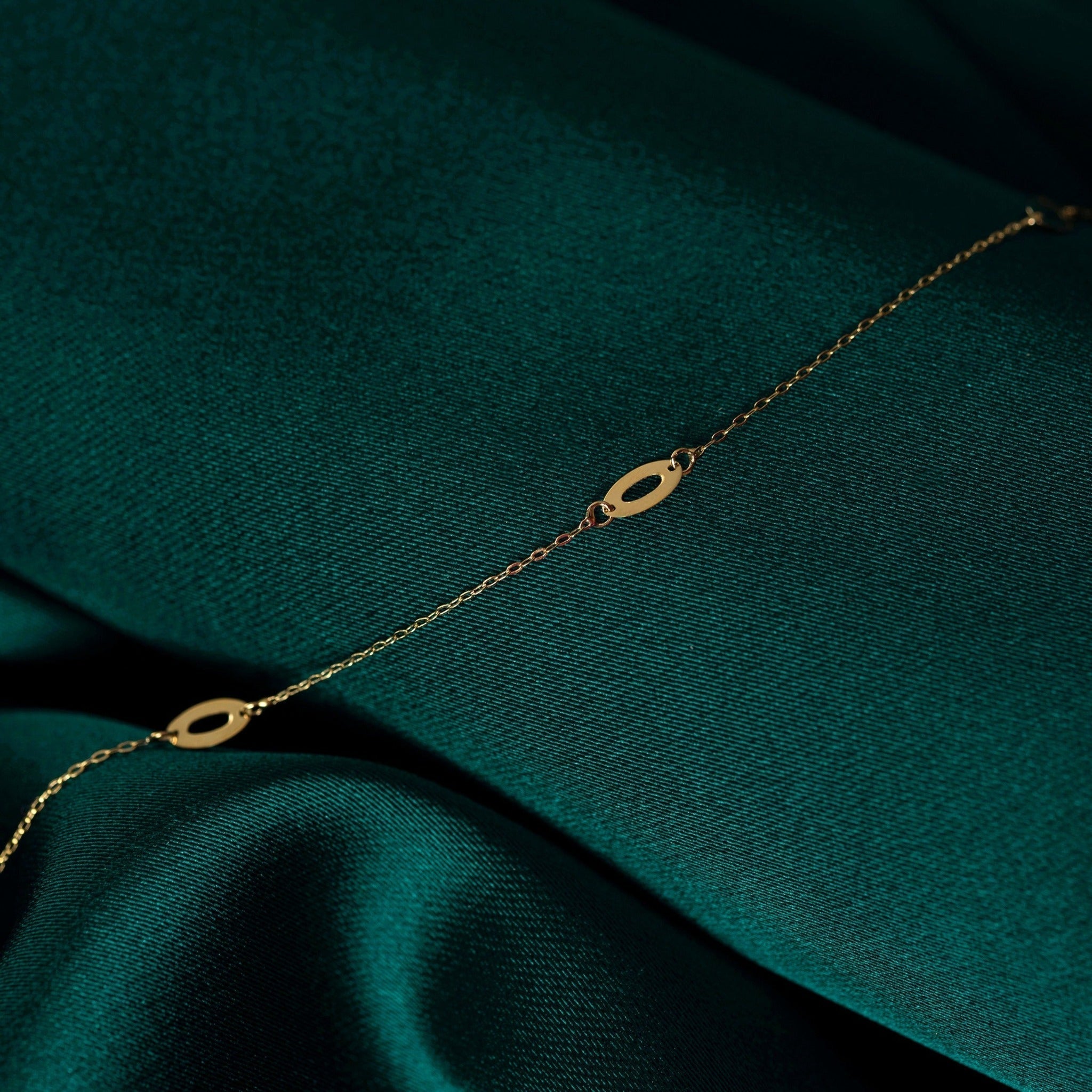Fine gold-toned chain bracelet with flat, open oval details, draped diagonally across dark teal fabric.