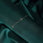 Fine gold-colored chain bracelet with two open flower motifs and small bead accents, draped diagonally over dark green fabric.